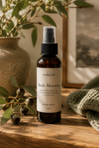 An amber room spray bottle labeled 'Sunday Folk Bush Morning' stands on a rustic wooden surface, surrounded by eucalyptus leaves, a textured ceramic vase, and a folded green knit blanket in warm natural light.