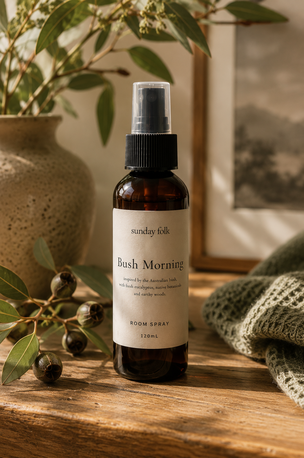 An amber room spray bottle labeled 'Sunday Folk Bush Morning' stands on a rustic wooden surface, surrounded by eucalyptus leaves, a textured ceramic vase, and a folded green knit blanket in warm natural light.