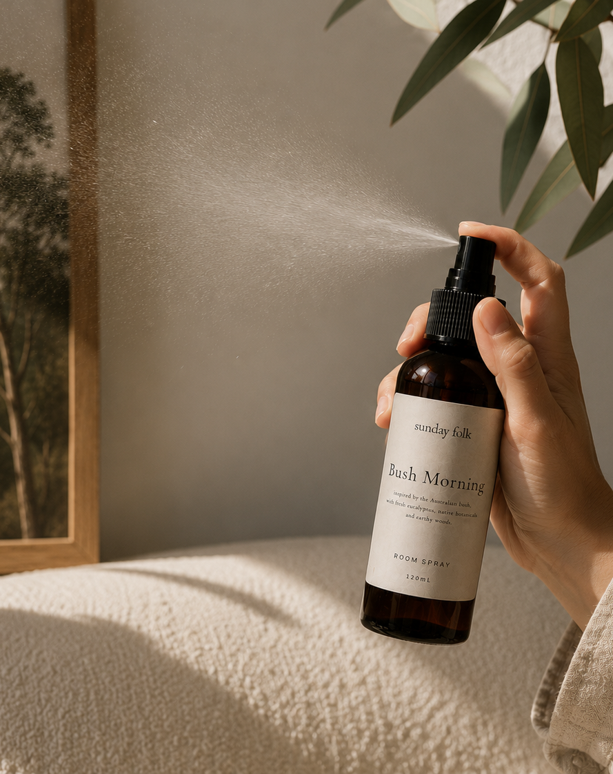 A hand holding a brown room spray bottle labeled 'Sunday Folk Bush Morning' as it sprays a fine mist. The scene is set in a warm, sunlit room with a textured cream blanket, soft shadows, and a leafy plant in the background.
