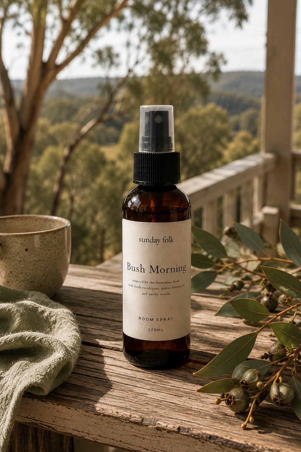 An amber room spray bottle labeled 'Sunday Silk Bush Morning' sits on a rustic wooden deck with a blurred outdoor landscape background, surrounded by a textured bowl, green cloth, and eucalyptus leaves.