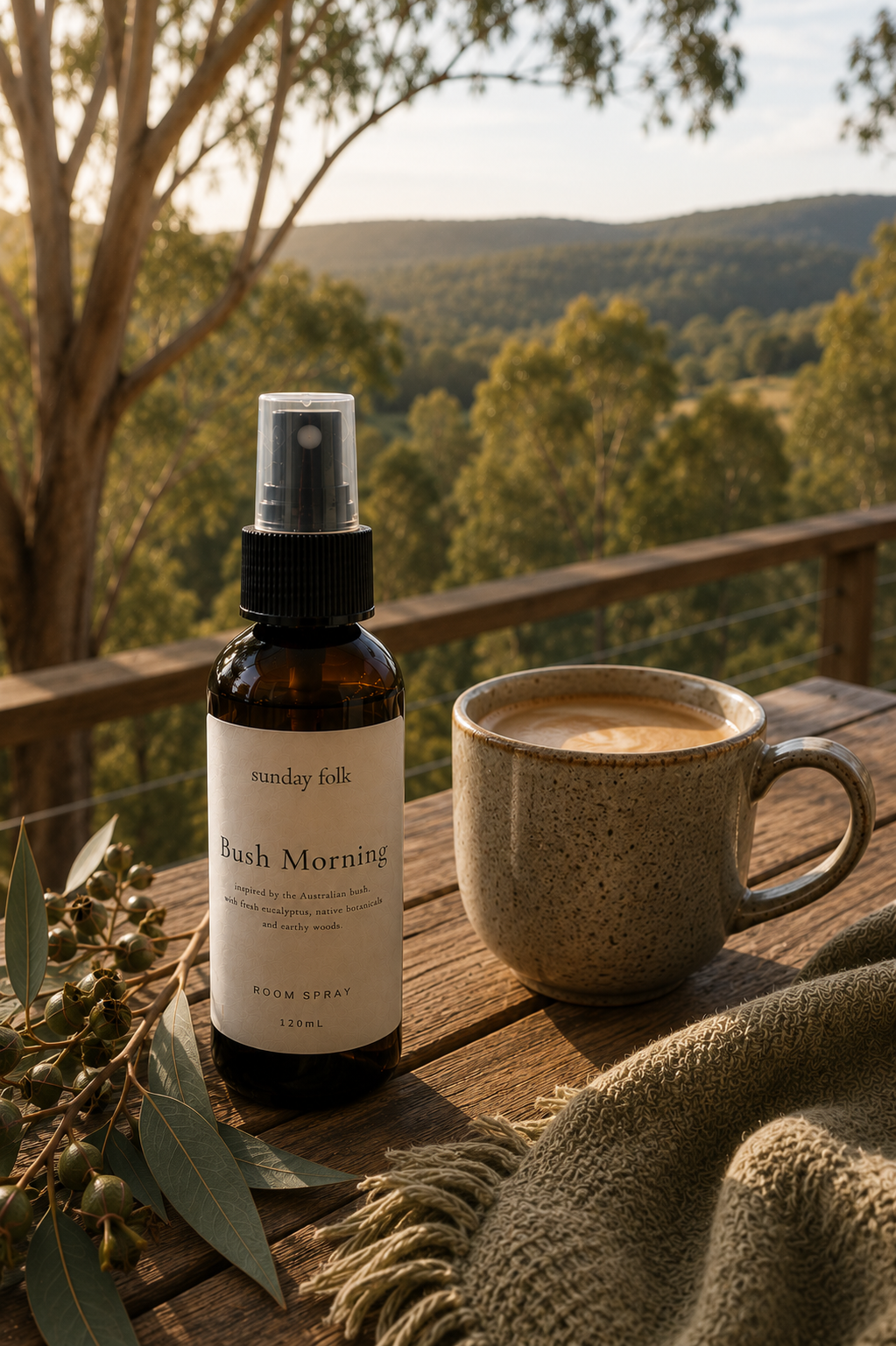 An amber spray bottle labeled 'Sunday Folk  Bush Morning' stands on a wooden table beside a ceramic mug filled with coffee, with a knitted throw and eucalyptus branches in the foreground and a scenic forested hillside in the background.